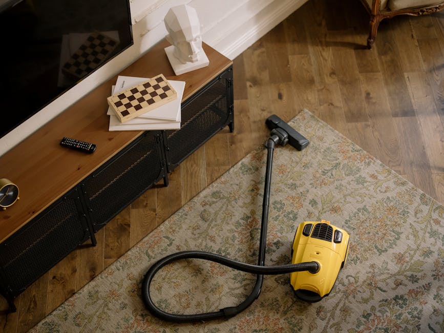 A yellow portable vacuum cleaner with a black hose attachment is situated on a patterned area rug in a living room corner. Nearby, there is a wooden sideboard with a chessboard, some stacked papers, a decorative bust, and a remote control on its surface. The room features wooden flooring, white baseboards, and a section of a chalkboard or dark wall visible in the background. The scene appears clean and well-maintained, illustrating a domestic cleaning process carried out by Little Ilford Carpet Cleaners as part of a surface cleaning or deep cleaning procedure.