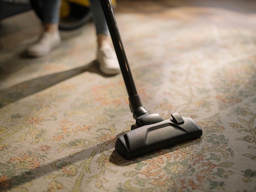 A yellow portable vacuum cleaner with a black hose attachment is situated on a patterned area rug in a living room corner. Nearby, there is a wooden sideboard with a chessboard, some stacked papers, a decorative bust, and a remote control on its surface. The room features wooden flooring, white baseboards, and a section of a chalkboard or dark wall visible in the background. The scene appears clean and well-maintained, illustrating a domestic cleaning process carried out by Little Ilford Carpet Cleaners as part of a surface cleaning or deep cleaning procedure.