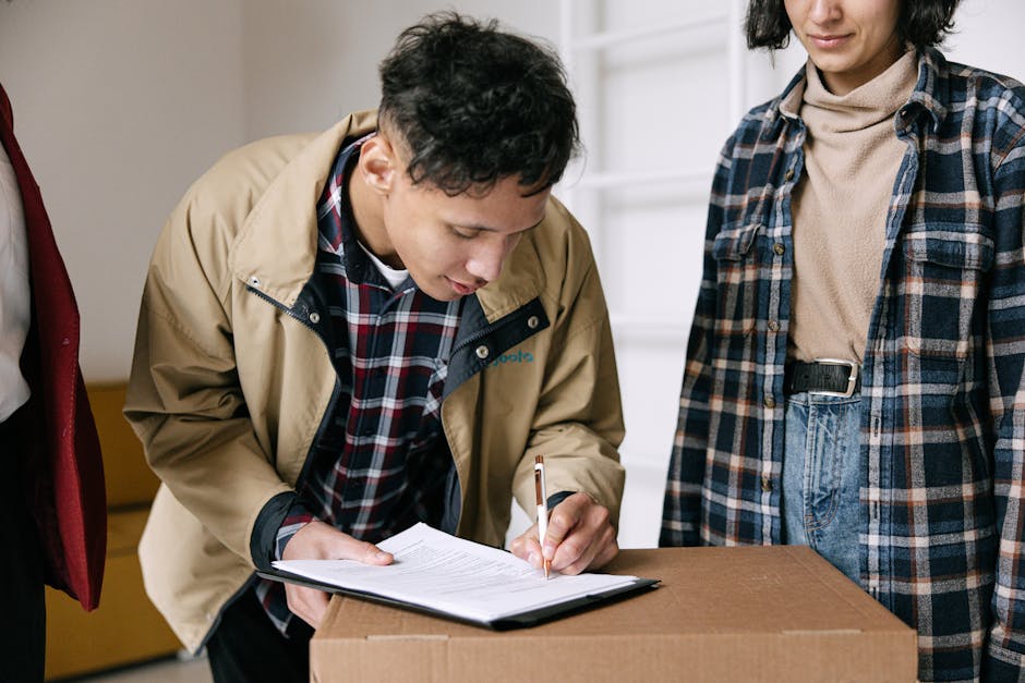 A man wearing a beige jacket and plaid shirt is signing a document placed on a cardboard box, while a woman dressed in a plaid shirt and beige turtleneck observes beside him. They are in a well-lit ro