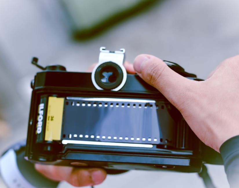 Close-up of a person holding a professional film camera, focusing on the camera's viewfinder and film reel at the top. The person’s hands are visible, with one hand gripping the side of the camera and